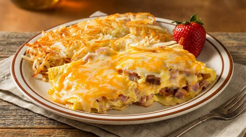 A plate of cheesy ham and cheese omelet with hash browns and a strawberry.