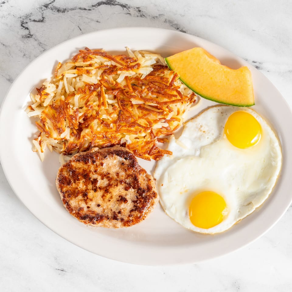 Plate of crispy hash browns, two sunny-side-up eggs, grilled sausage patty, and cantaloupe wedge on a white plate.