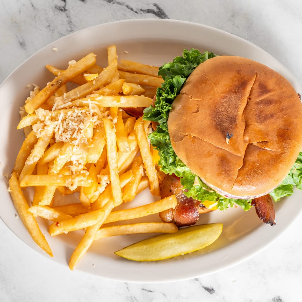 Bacon cheeseburger with lettuce, cheese-topped fries, and a pickle spear on a white plate.