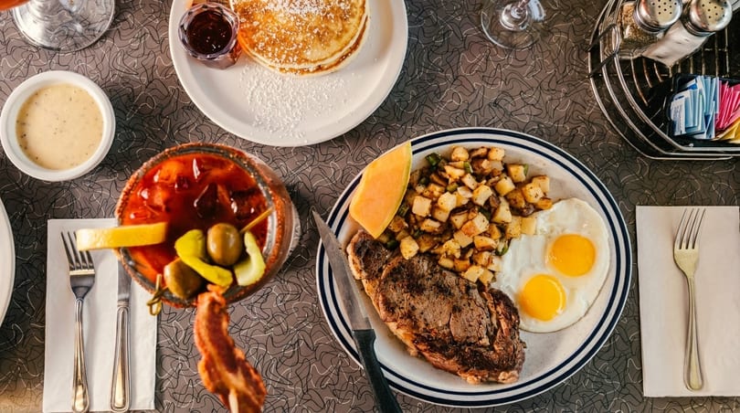 Overhead view of a diner breakfast with steak, eggs, potatoes, pancakes, and a Bloody Mary.