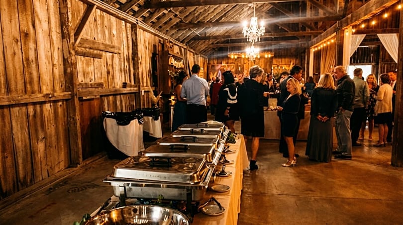 A rustic barn interior with a buffet line and guests mingling under chandeliers and string lights.