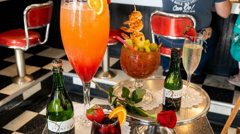 Assortment of colorful cocktails and champagne bottles on a silver tray with red diner stools in the background.