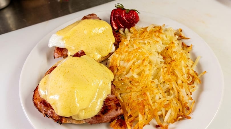 Eggs Benedict with hollandaise sauce, hash browns, and a strawberry on a white plate.