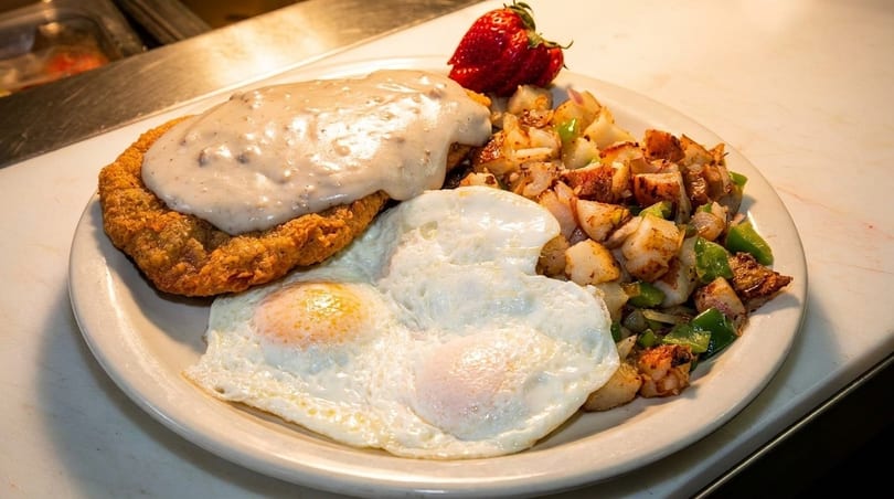 Chicken fried steak with gravy, two fried eggs, home fries, and a strawberry on a white plate.