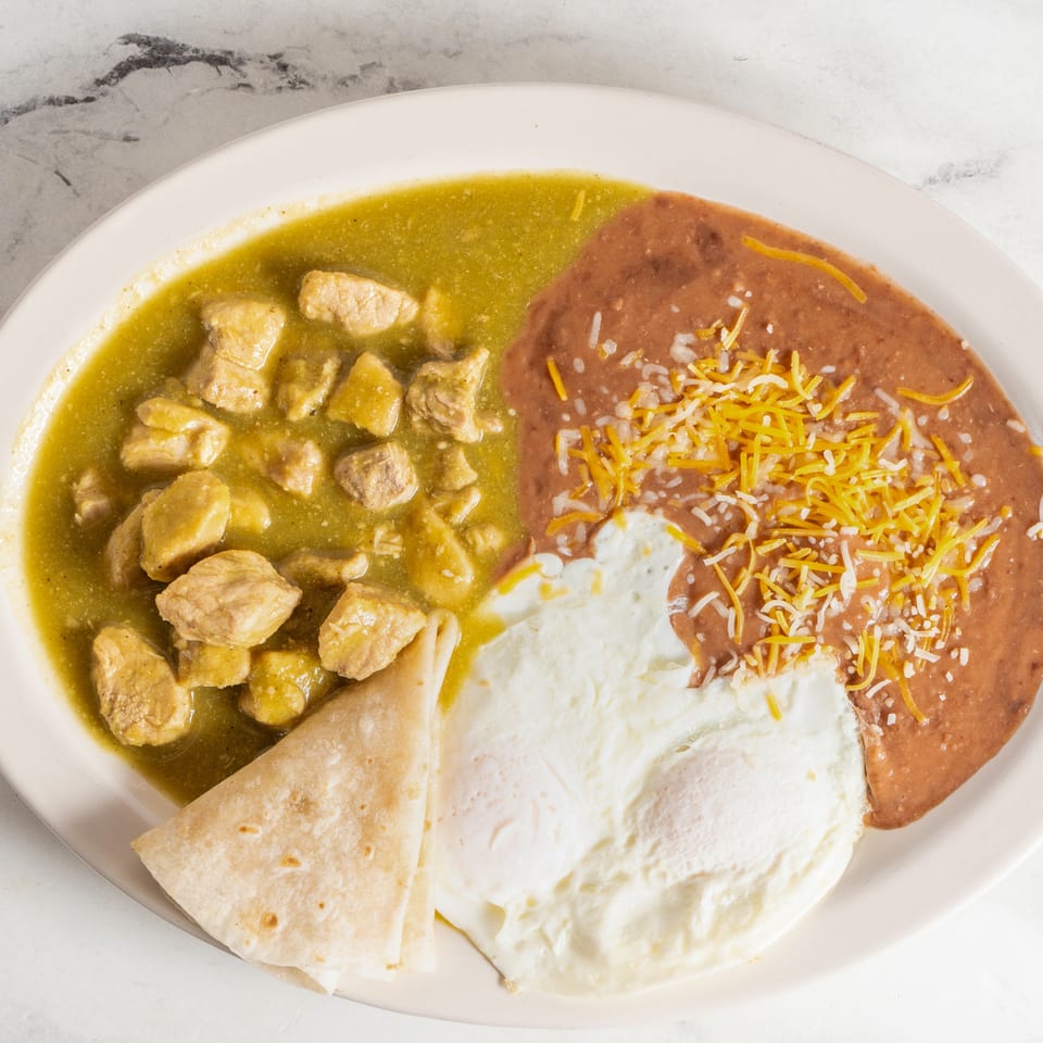 Mexican breakfast plate with pork in green chile sauce, refried beans topped with cheese, fried eggs, and flour tortillas.