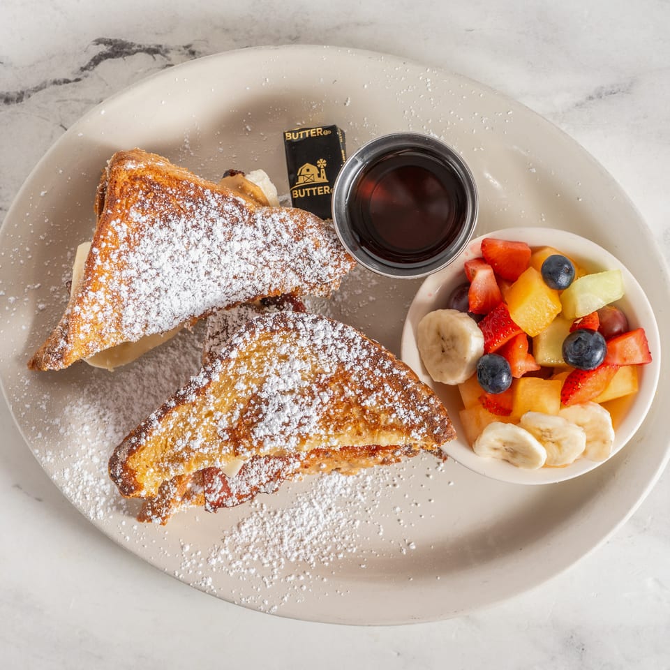 Powdered sugar-dusted French toast with syrup, butter, and a bowl of mixed fresh fruit on a plate.