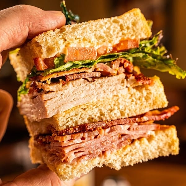 A hand holds a thick club sandwich with turkey, ham, bacon, lettuce, and tomato.