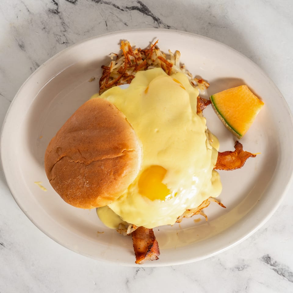 Breakfast plate with eggs Benedict, crispy bacon, hash browns, toasted bun, and cantaloupe wedge on a marble table.