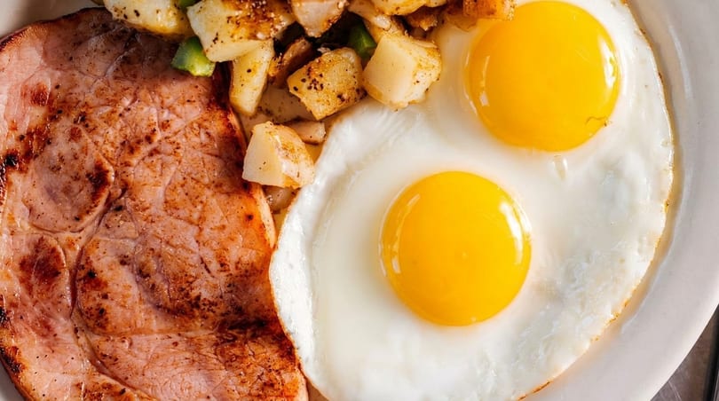 Close-up of a breakfast plate with grilled ham, crispy home fries, and two sunny-side-up eggs.