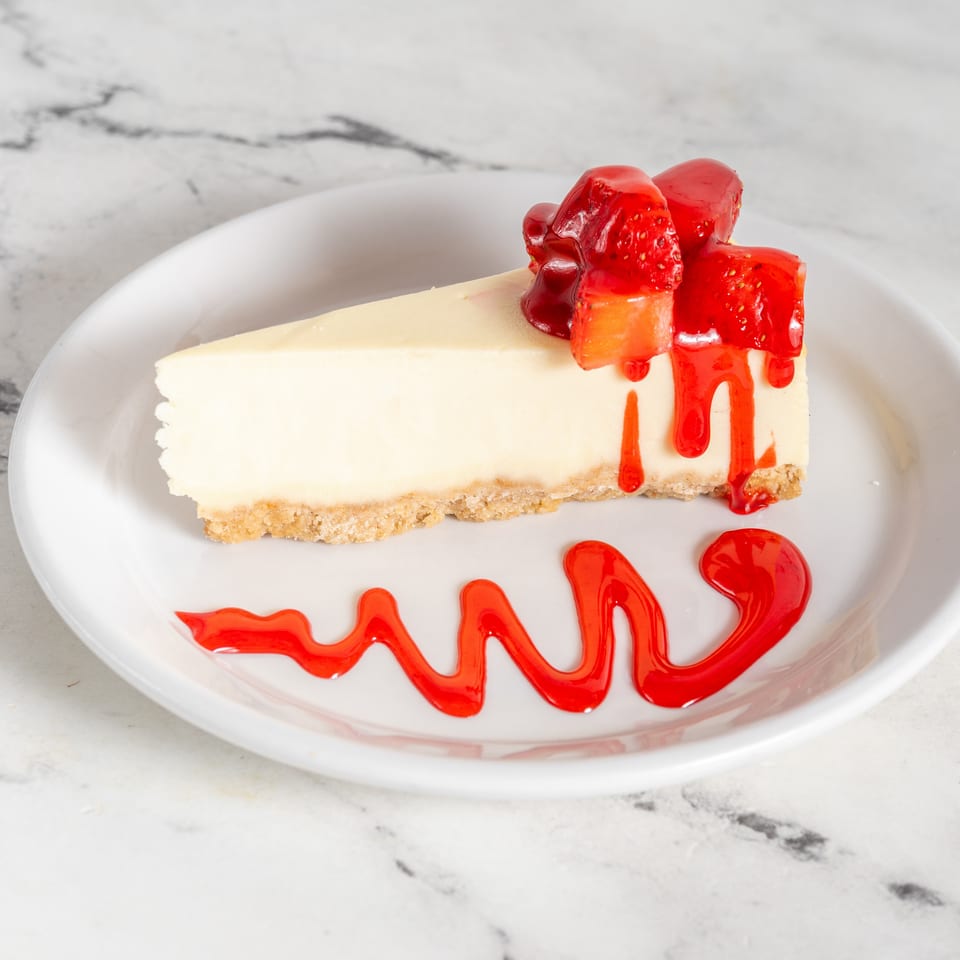 Slice of creamy cheesecake topped with strawberries and red berry sauce on a white plate.
