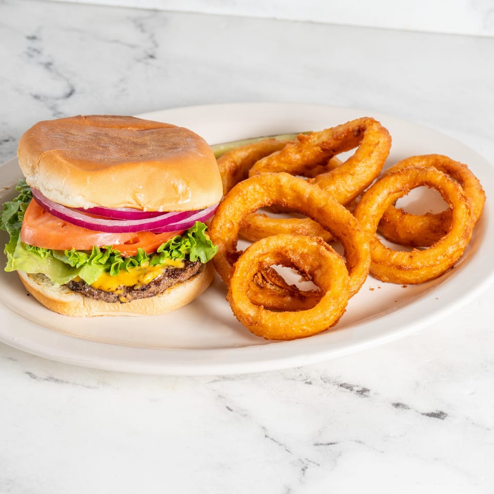 Cheeseburger topped with lettuce, tomato, and red onion served alongside crispy onion rings on a white plate.