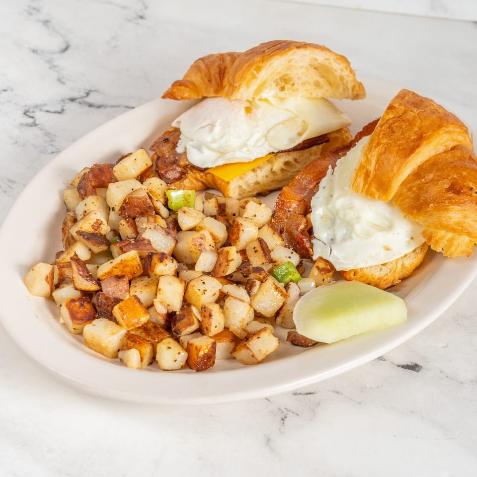 Breakfast plate with two croissant sandwiches containing eggs, cheese, and bacon, served with diced potatoes and melon.