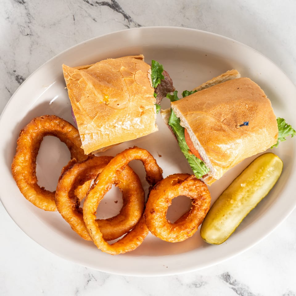 Two baguette sandwich halves with lettuce and tomato, crispy onion rings, and a pickle spear on a plate.