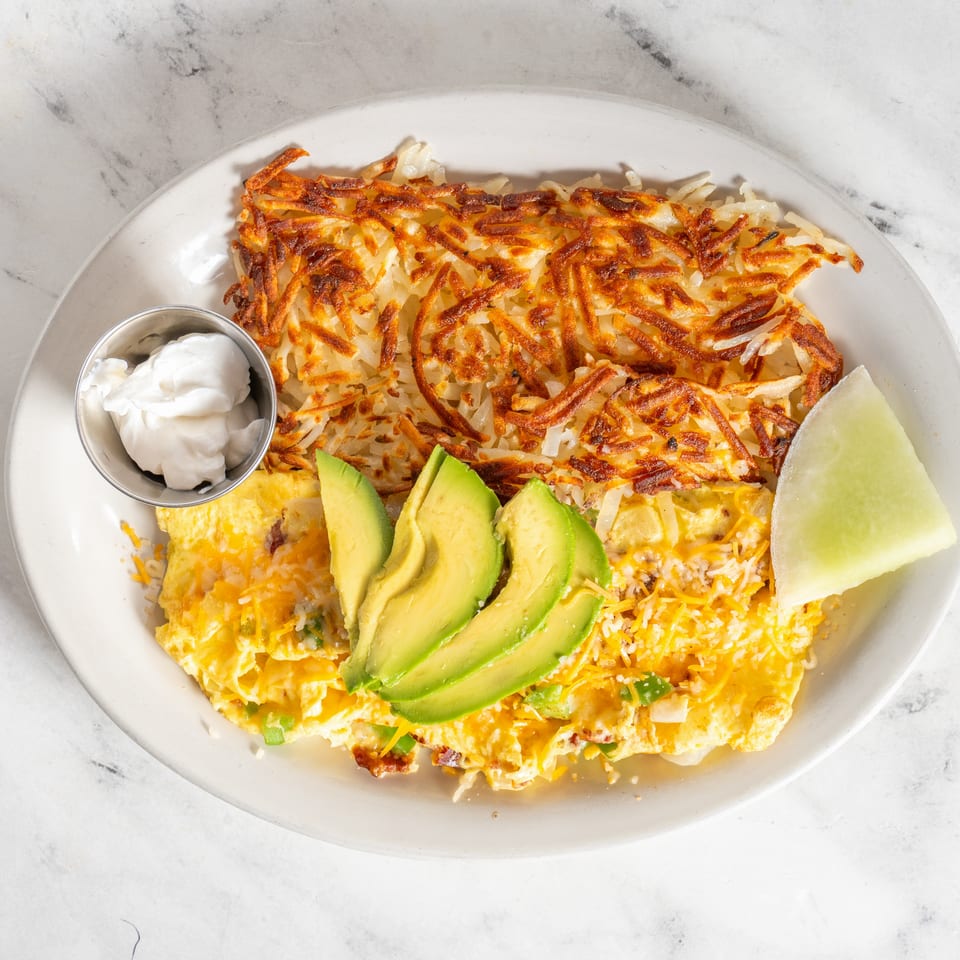 Cheesy omelet topped with avocado and crispy hash browns, served with sour cream and a lime wedge on a white plate.