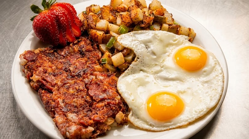 A breakfast plate with two fried eggs, corned beef hash, home fries, and a strawberry.