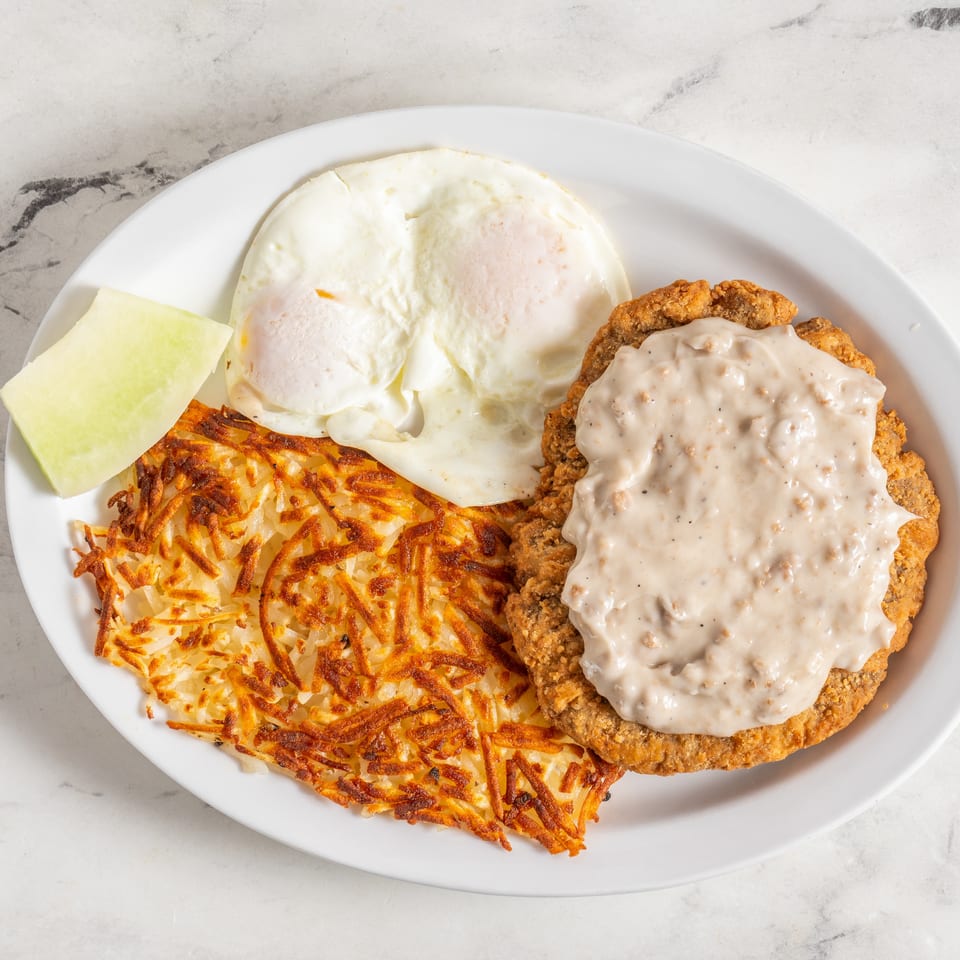 Plate of crispy fried chicken topped with creamy gravy, two fried eggs, golden hash browns, and melon.