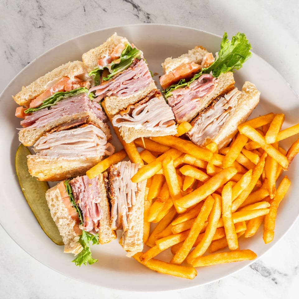 Club sandwich quarters with turkey, ham, lettuce, tomato, and mayonnaise served alongside French fries and a pickle.