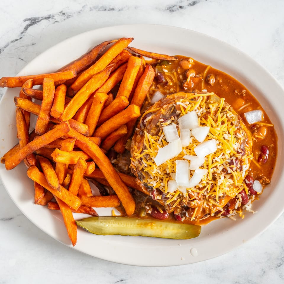 Plate of sweet potato fries beside a chili-topped baked potato with cheese, onions, beans, and a pickle spear.