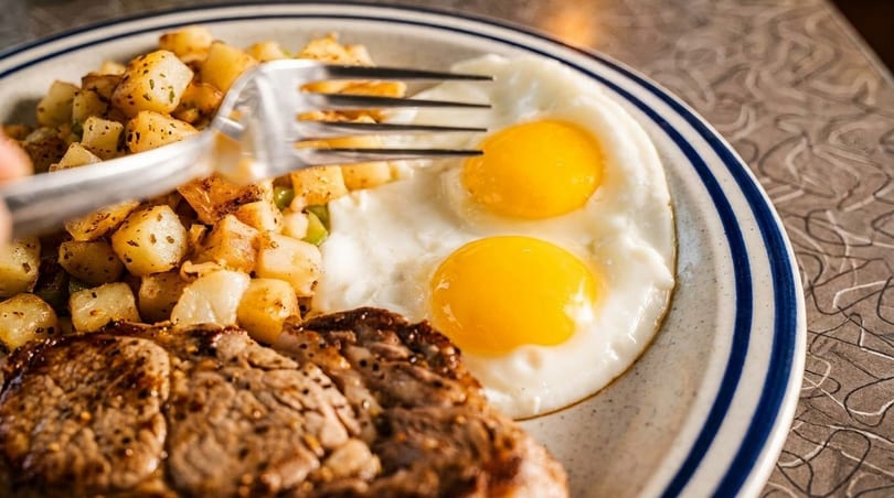 Steak and eggs breakfast with two sunny-side-up eggs, seasoned potatoes, and a fork on a diner plate.