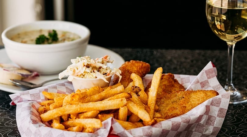 Fish and chips with coleslaw in a basket, a bowl of soup, and a glass of white wine.