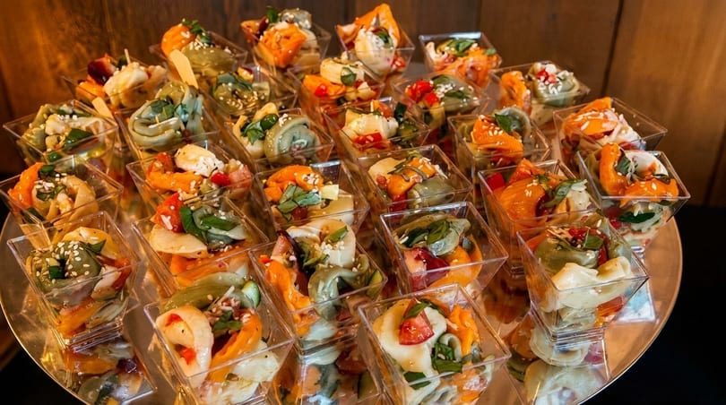 Numerous small clear cups filled with colorful pasta salad, garnished with herbs and sesame seeds.