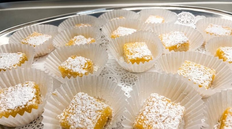 A silver tray filled with individual lemon bars, each dusted with powdered sugar in white paper cups.