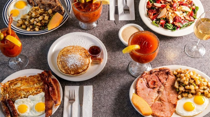 A table laden with various breakfast dishes, including eggs, bacon, pancakes, and Bloody Marys.