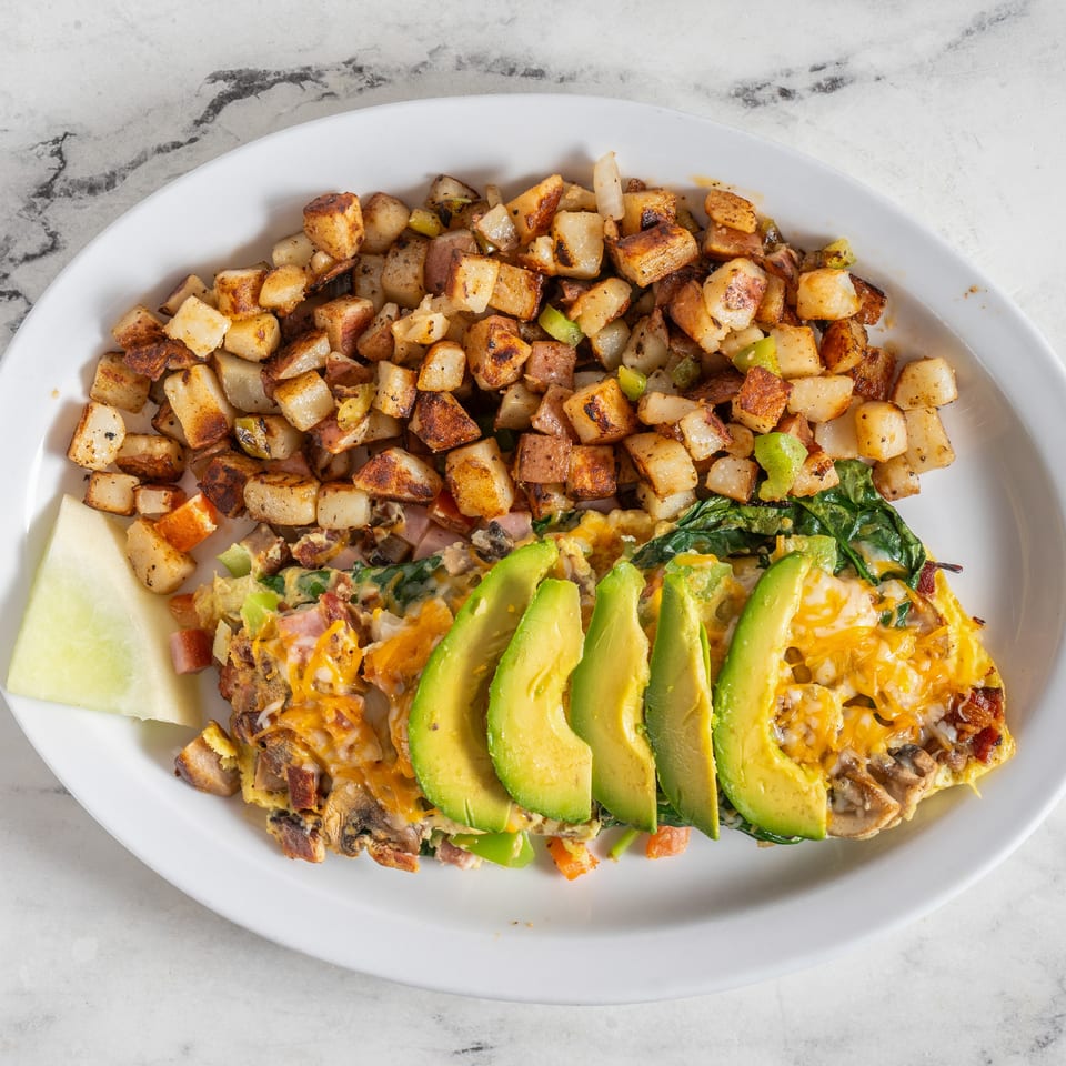 Breakfast plate with cheesy vegetable-and-bacon omelet, sliced avocado, roasted potatoes, and melon on a white platter.