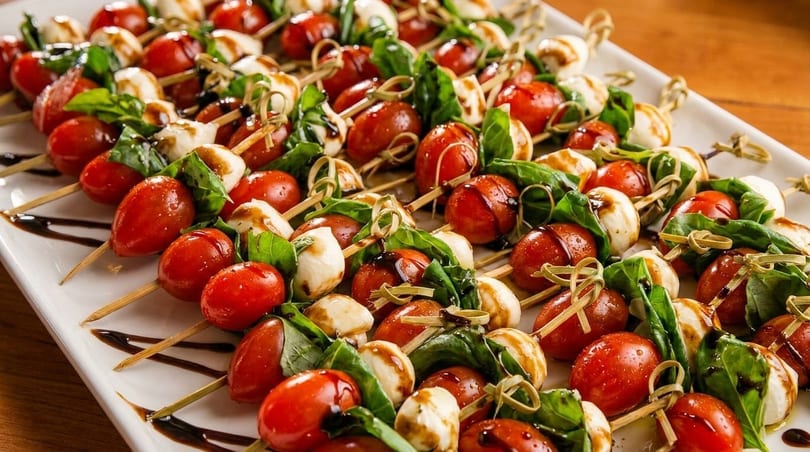 Caprese skewers with cherry tomatoes, mozzarella, basil, and balsamic glaze on a white platter.