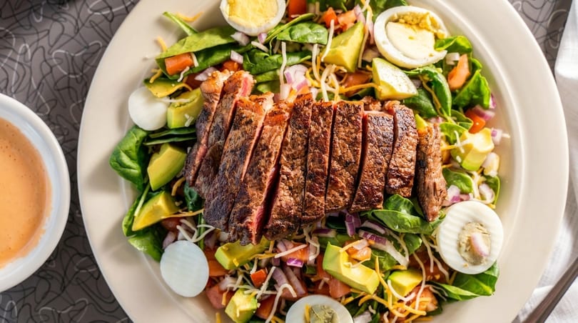 Overhead view of a steak salad with avocado, spinach, tomatoes, eggs, and cheese.