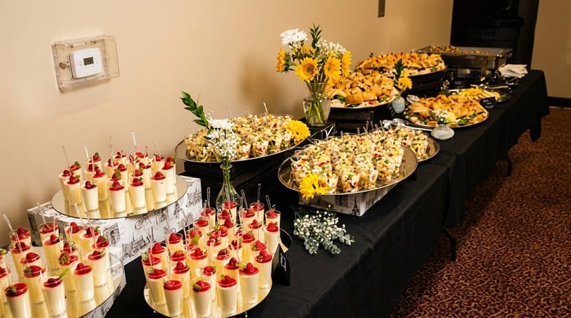 A buffet table laden with desserts, appetizers, and sandwiches, adorned with sunflowers and white flowers.