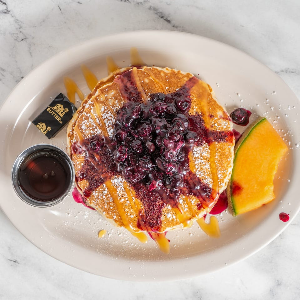 Stack of pancakes topped with blueberries, caramel drizzle, powdered sugar, and melon, served with syrup and butter.