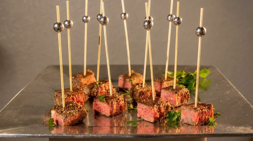 Seared beef cubes on skewers with silver ball tops, garnished with parsley on a reflective tray.