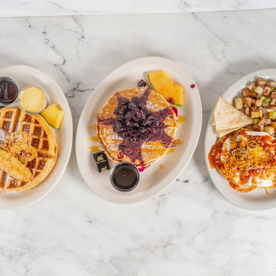 Overhead view of waffles with fried chicken, blueberry pancakes, and a cheesy breakfast plate on white marble.
