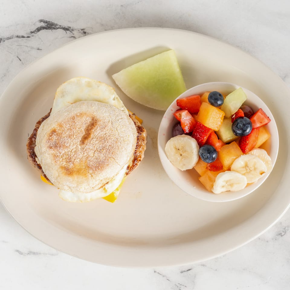 Breakfast plate with an egg and cheese sandwich, melon wedge, and bowl of mixed fruit including berries, banana, and strawberries.