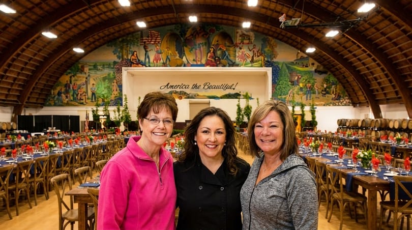 Three women smiling in a large hall with set tables, wine barrels, and a mural.