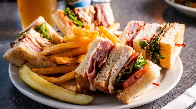 Club sandwiches with fries and a pickle spear on a white plate, with a beer in the background.
