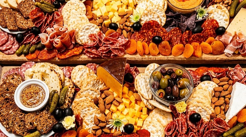 Two large charcuterie boards overflowing with meats, cheeses, crackers, olives, nuts, and dried apricots.