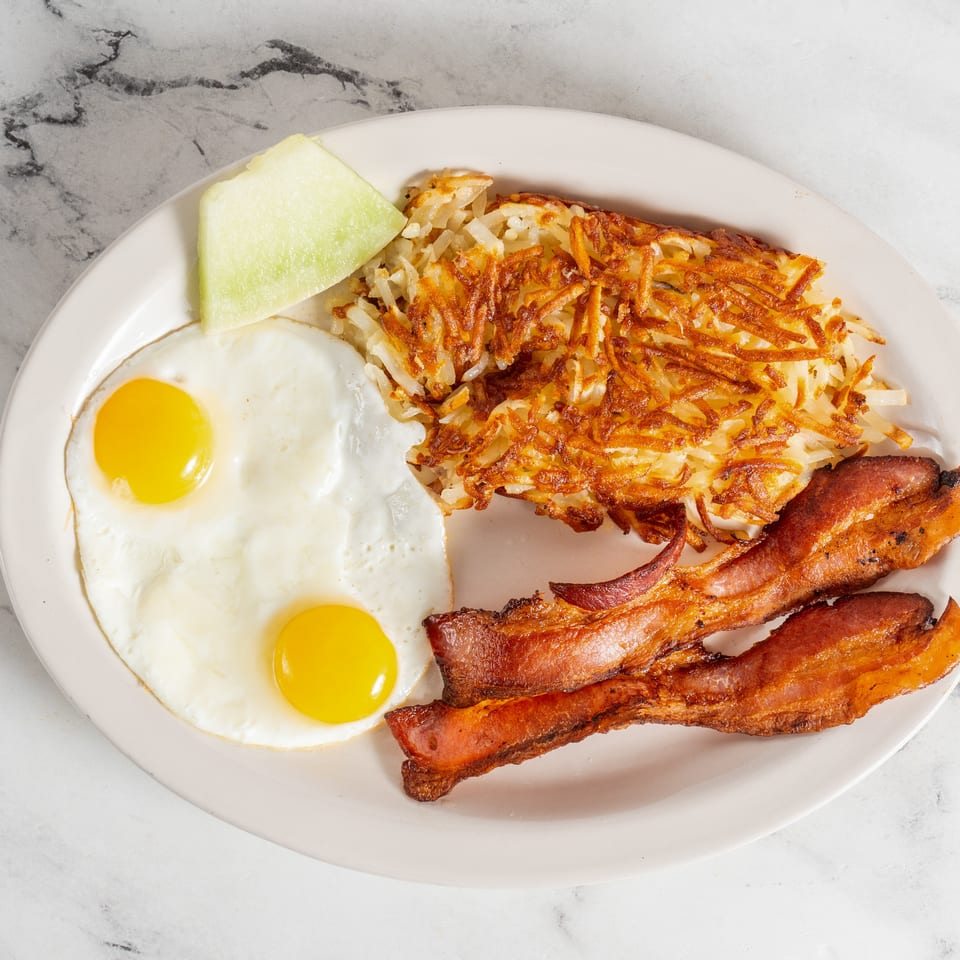 Breakfast plate with two sunny-side-up eggs, crispy hash browns, bacon strips, and a slice of melon.