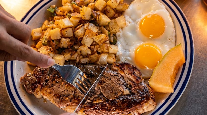 A person cutting into a steak with fried eggs, potatoes, and a cantaloupe slice on a plate.