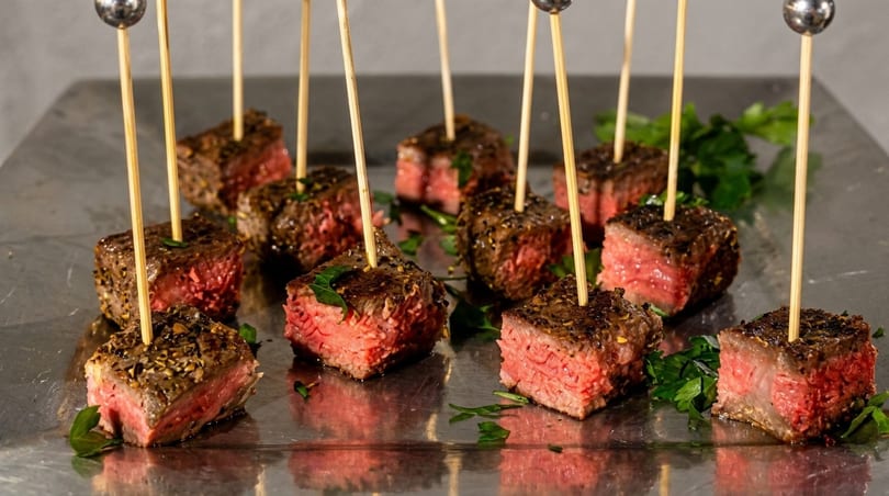 Close-up of numerous medium-rare steak bites on skewers, garnished with parsley on a metallic tray.