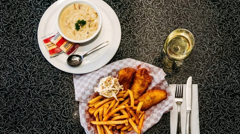 Clam chowder, fish and chips with coleslaw, and a glass of white wine on a diner table.