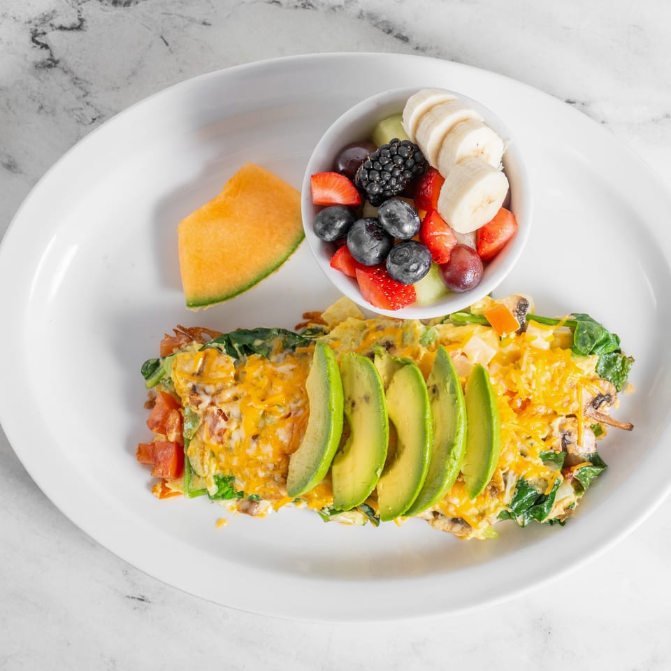 Cheesy vegetable omelet topped with sliced avocado, served with a bowl of mixed berries, banana, and cantaloupe.