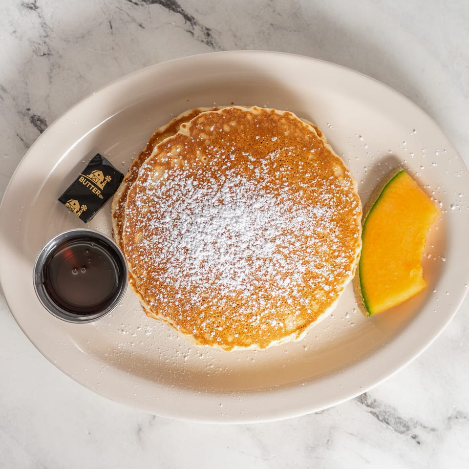 Stack of pancakes dusted with powdered sugar, served with syrup, butter, and cantaloupe on a plate.