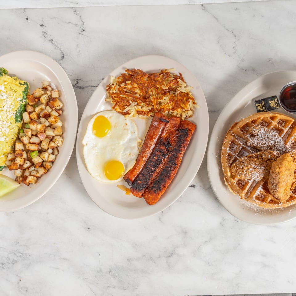 Overhead view of three breakfast plates featuring spinach-and-feta omelet, eggs with bacon and hash browns, and powdered-sugar waffles with chicken.