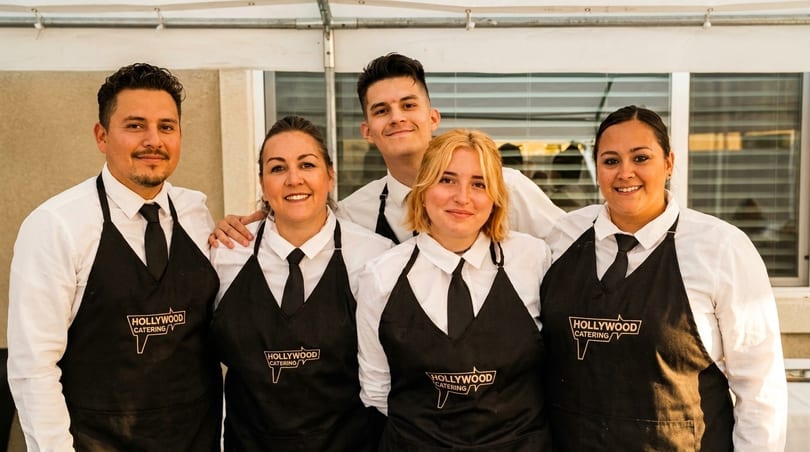 Five smiling Hollywood Catering staff members in black aprons and white shirts pose for a photo.