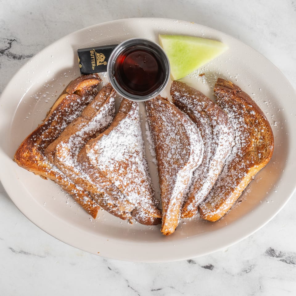 French toast triangles dusted with powdered sugar, served with syrup, butter, and melon on a plate.