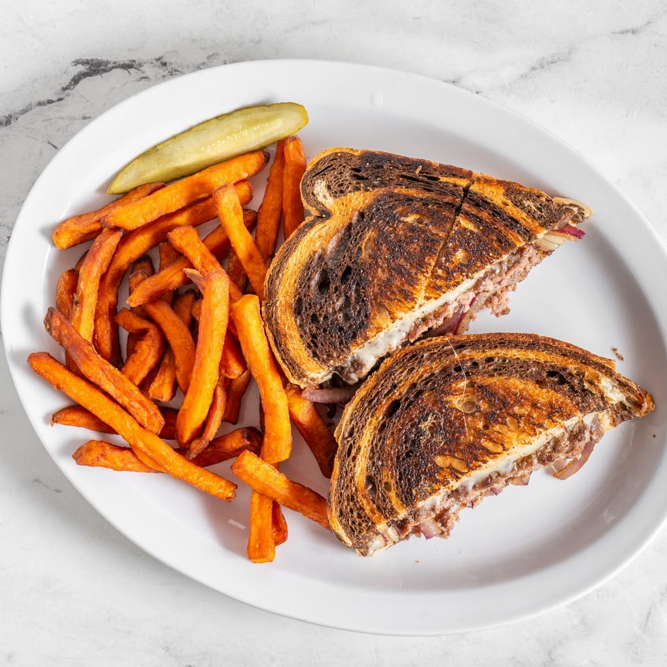 Grilled pastrami sandwiches with melted cheese, sweet potato fries, and a pickle spear on a white plate.