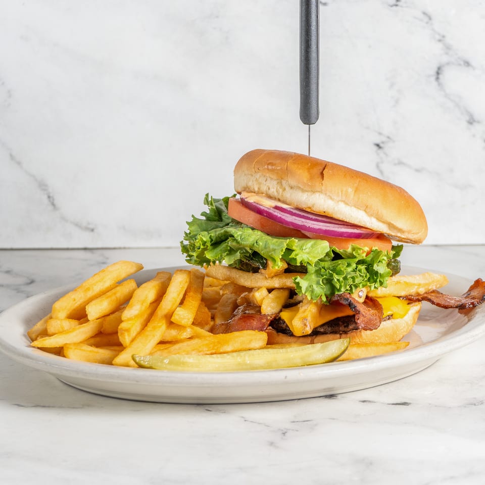 Bacon cheeseburger with lettuce, tomato, onion, and pickles served with French fries on a white plate.