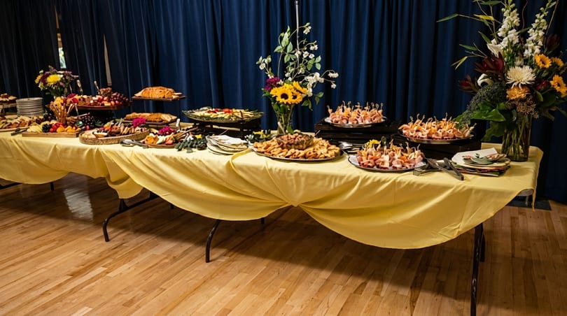 A long buffet table with a yellow tablecloth, laden with appetizers and floral arrangements.